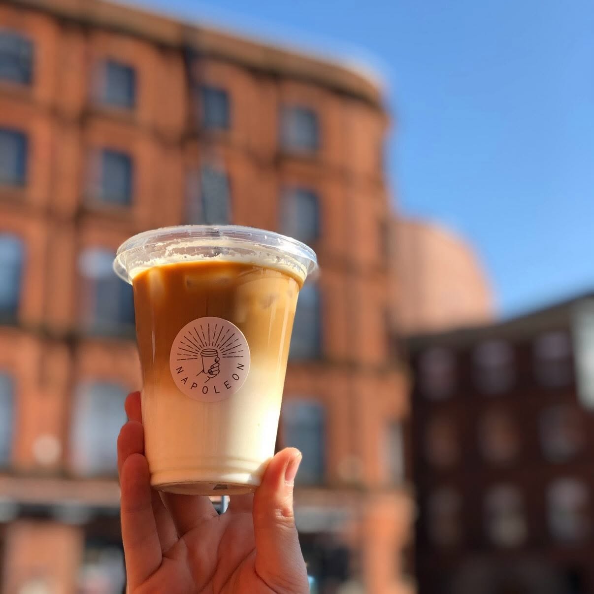 An iced latte held up against red-brick Belfast buildings and blue sky